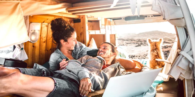 Couple relaxing on the bed in a campervan with the side door open and a dog looking out