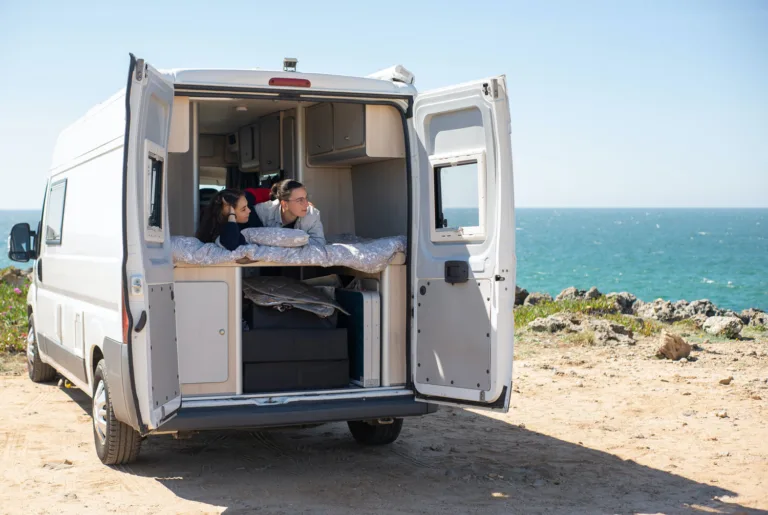 Couple lying on the bed of a white panel van conversion with the rear doors open to the sea
