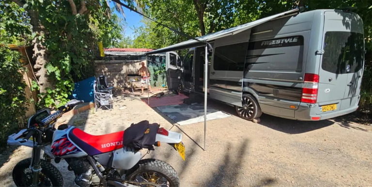 Julie's grey Mercedes Sprinter with its awning out on a campsite, a Honda trail bike parked in front