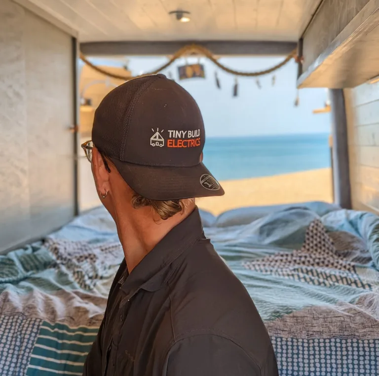 Man in a Tiny Build Electrics cap looking out of the open back of a van at the beach