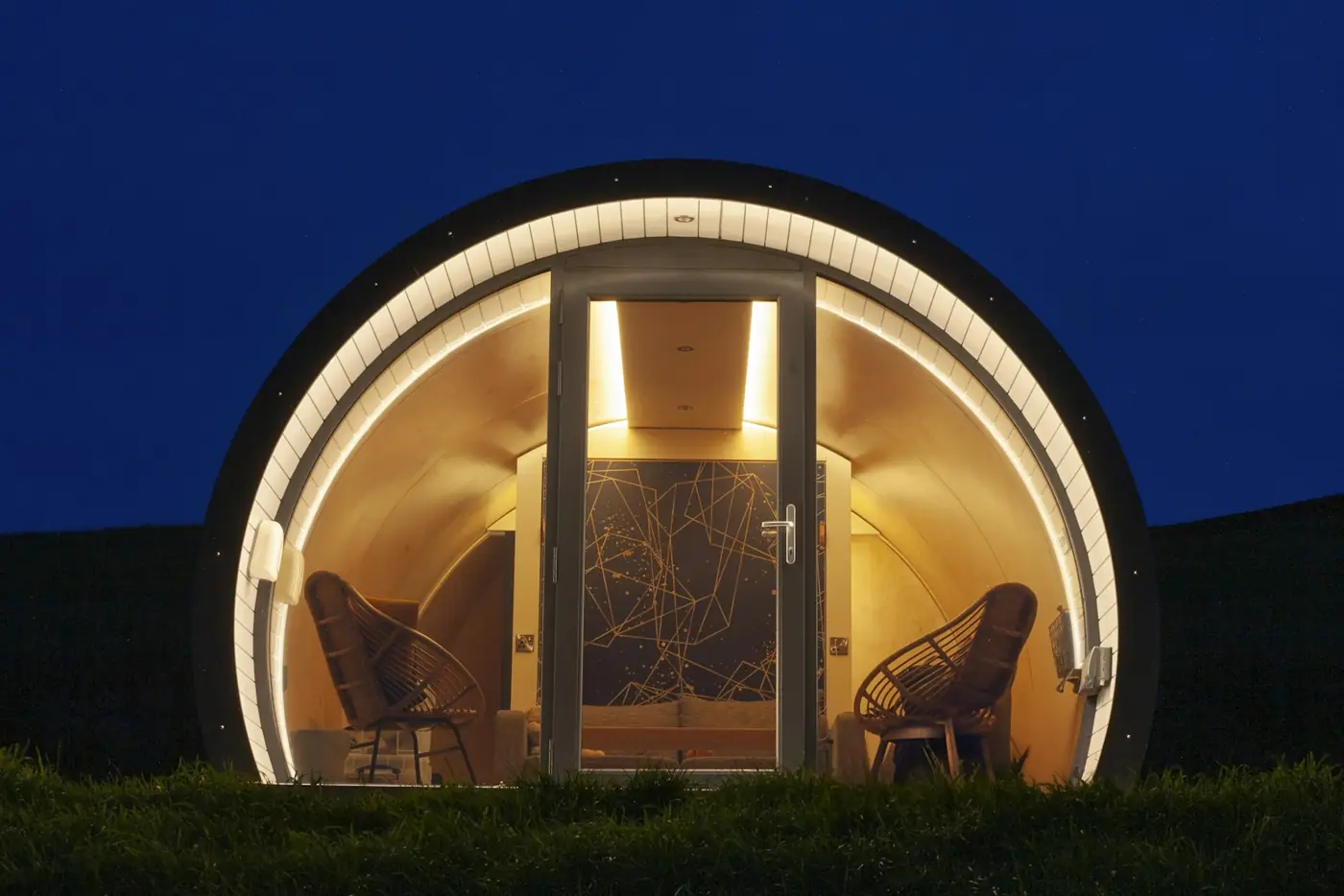 Arched glamping pod lit warmly from inside at night, two chairs behind the glass door