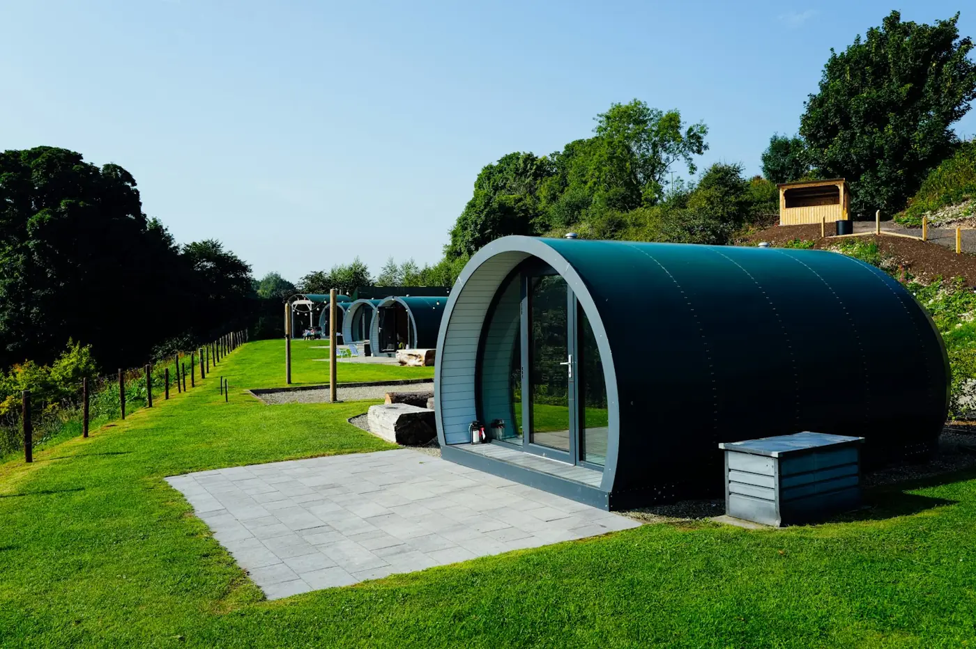 Row of green arched glamping pods on level grass pitches with paved patios on a rural glamping site