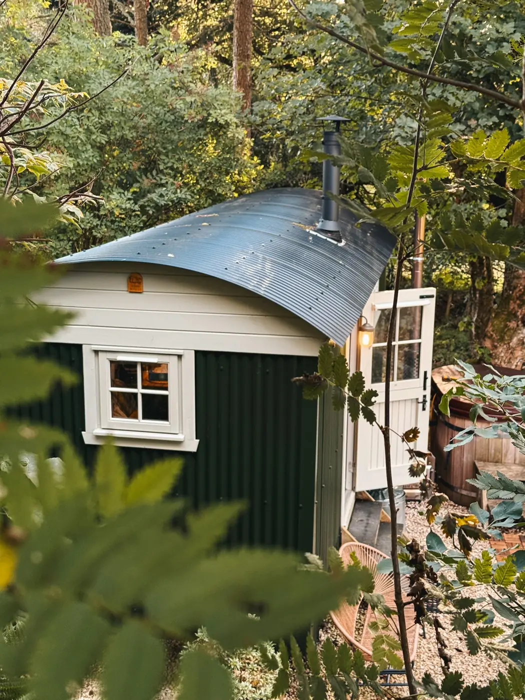 Green shepherd's hut with a curved metal roof and wood-burner flue among trees