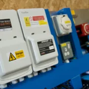 Two FuseBox consumer units with Tiny Build Electrics and Danger 230 Volts labels, a dual-supply warning label and an inspection notice, mounted beside a hook-up inlet, a socket, a battery isolator and Victron equipment on a blue test rig