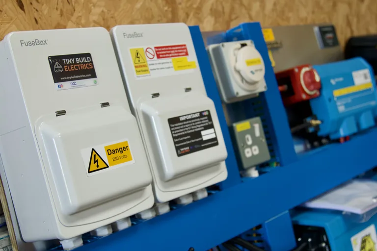 Two FuseBox consumer units with Tiny Build Electrics and Danger 230 Volts labels, a dual-supply warning label and an inspection notice, mounted beside a hook-up inlet, a socket, a battery isolator and Victron equipment on a blue test rig