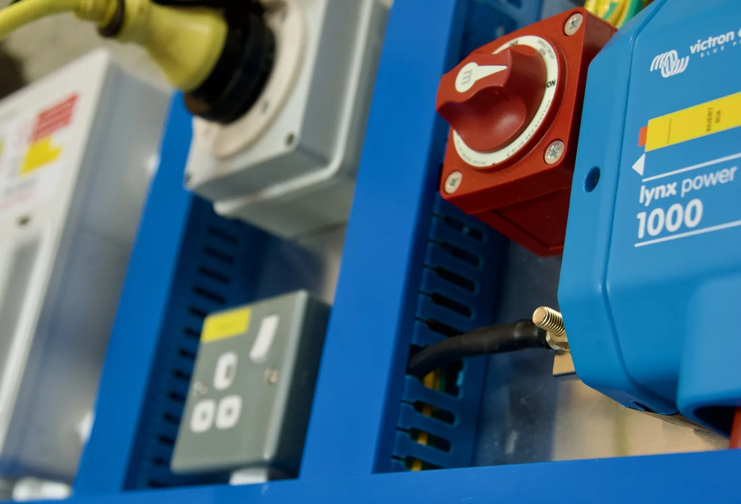 Close-up of a Tiny Build Electrics board in blue slotted trunking with a Victron Lynx Power In, a red battery switch, a hook-up inlet and a 230V socket