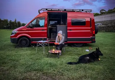 Red VW Crafter campervan with the side door open, a fire pit and a dog on the grass at dusk