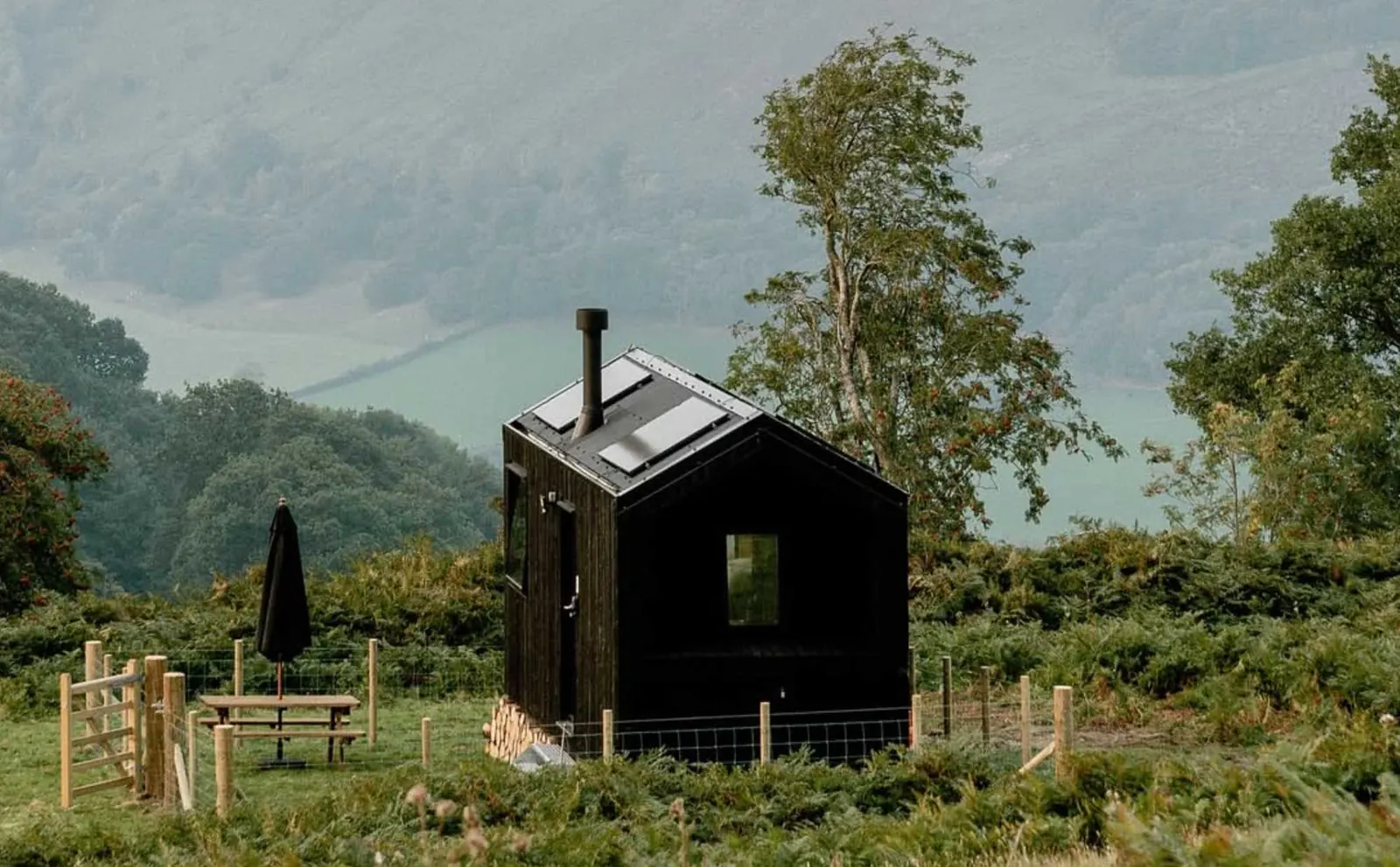 Small black timber off grid cabin with an off grid solar system on the roof and a wood burner flue, on a fenced hillside above a misty UK valley
