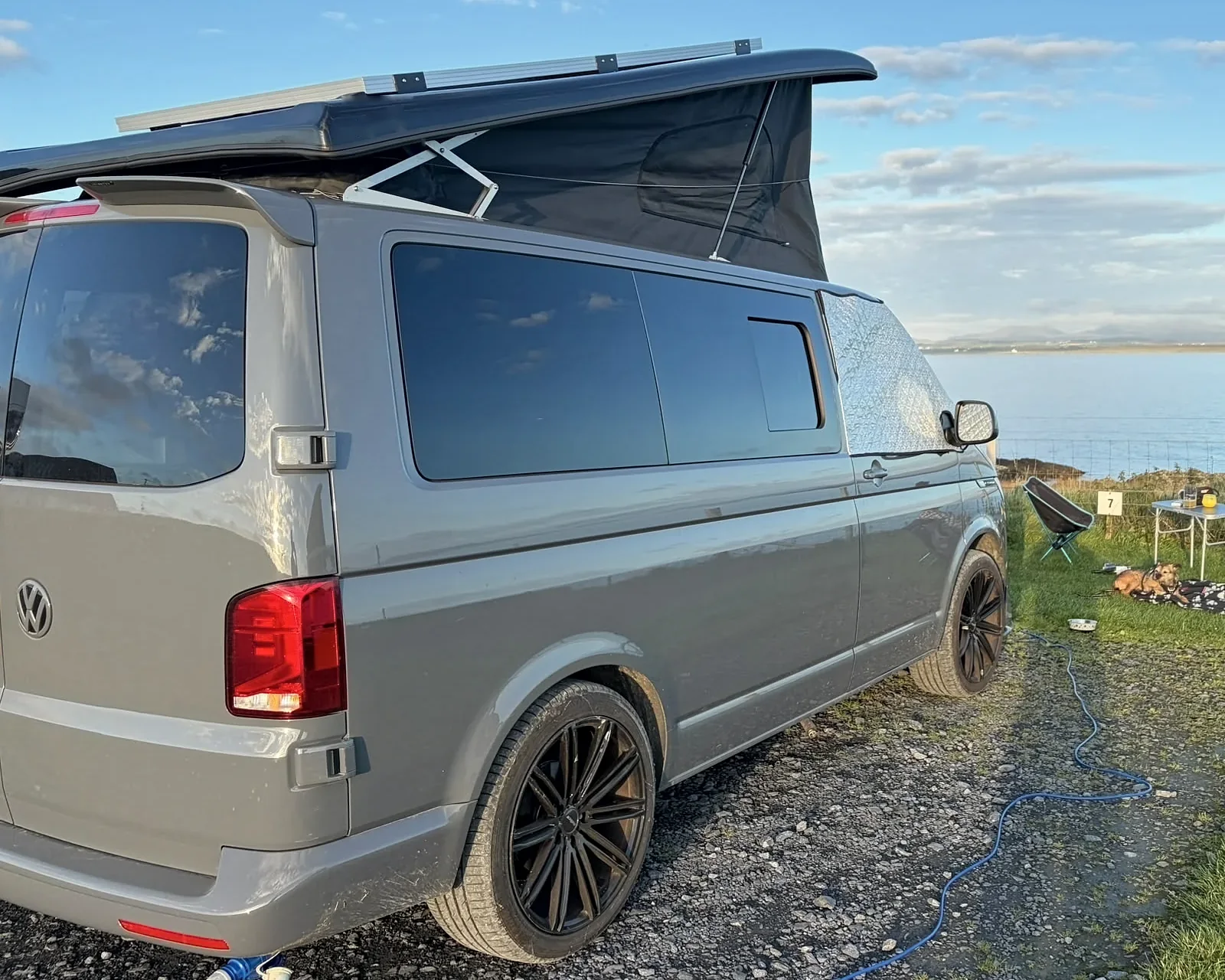 Grey VW T6.1 campervan parked side-on above the sea, with its pop-top roof raised and tinted windows along the side
